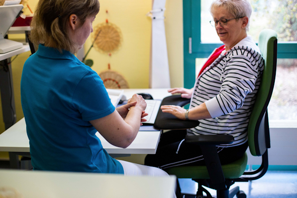 Therapeutin erklärt einer am Schreibtisch sitzenden Rehabilitandin die ergonomische Handhaltung 